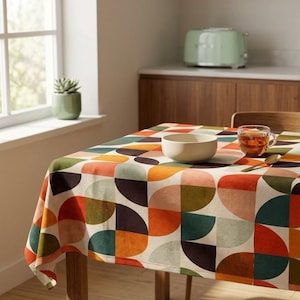 Puede incluir: Un mantel colorido con un patrón geométrico de formas naranjas, verdes y marrones. Un cuenco blanco, un vaso de té y una cuchara están sobre la mesa. Una tostadora verde es visible en el fondo.