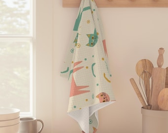 May include: A cream-colored kitchen towel with a repeating pattern of stylized cats, trees, and geometric shapes in teal, coral, and yellow. The towel is hanging on a wooden rack with pegs, suggesting a kitchen setting.