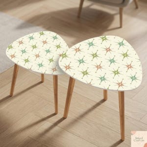 May include: Two small, cream-colored side tables with a starburst pattern in shades of green, orange, and blue. The tables have a triangular shape and are supported by three wooden legs. The tables are placed on a light-colored wooden floor.