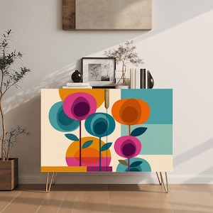 May include: A rectangular cabinet featuring a colorful graphic print of stylized flowers in shades of magenta, orange, and teal on a cream background. The piece is supported by four thin, gold-toned metal hairpin legs and has a flat top surface.