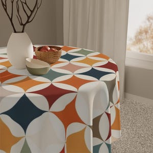 May include: A round tablecloth with a retro geometric pattern in shades of orange, blue, red, and green. A white vase, a small bowl, and a basket of apples sit on the table. The tablecloth is made of a soft, durable fabric.