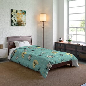 Op de afbeelding: Een turquoise dekbed met een modern atomisch ontwerp uit het midden van de eeuw. Het patroon bevat atoomvormen, sterren en gele en zwarte abstracte vormen, met witte lijnen op een turquoise achtergrond. Het dekbed ligt op een donkerbruin houten bedframe.