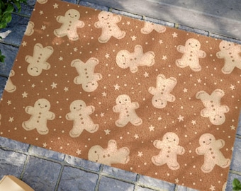 May include: A rectangular doormat featuring a brown background adorned with a repeating pattern of gingerbread men and white stars. The gingerbread men have smiling faces and are in various poses. The mat is placed on a blue stone surface.