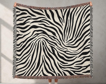 Könnte beinhalten: Eine große, rechteckige Wurkdecke mit einem auffälligen Zebrastreifenmuster in Schwarz und Creme. Die Decke hat einen Fransenrand und wird vor einem neutralen Hintergrund gehalten. Das Design ist ein klassischer Animal-Print.