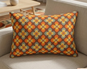 Op de afbeelding: Rechthoekig decoratief kussen met een retro bloemenpatroon. Het kussen heeft een bruine achtergrond met herhaalde bloemmotieven in oranje, geel en blauw. Het ligt op een neutraal gekleurde bank.