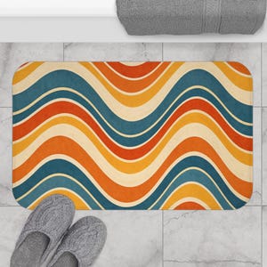 Puede incluir: Una alfombra de baño rectangular con un patrón de ondas retro en tonos azul, naranja, amarillo y crema. La alfombra está colocada sobre un suelo embaldosado, con un par de zapatillas grises en la parte inferior.