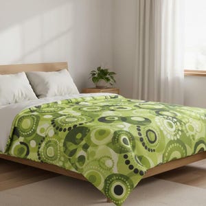 Puede incluir: Una manta estampada en verde y blanco sobre una estructura de cama de madera. La manta presenta un diseño repetitivo de círculos en varios tonos de verde, blanco y negro. Dos almohadas blancas descansan sobre la cama.