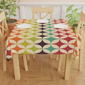 Könnte beinhalten: Ein farbenfrohes Tischtuch mit einem Retro-Geometrie-Muster. Das Design zeigt sich überlappende Diamantenformen in den Farbtönen Orange, Gelb, Grün, Türkis, Rot und Braun vor einem cremefarbenen Hintergrund. Vier weiße Teller liegen auf dem Tisch.