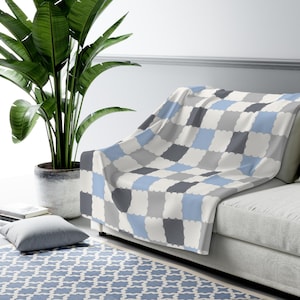 May include: A soft throw blanket featuring a repeating pattern of scalloped-edge squares in shades of light blue, dark gray, light gray, and off-white. The blanket is draped over the corner of a sofa, displaying a consistent grid design.