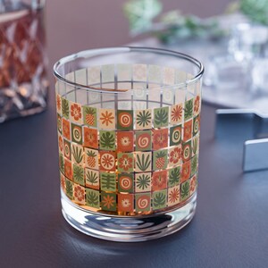 May include: A clear glass tumbler with a decorative pattern of colourful floral and geometric shapes. The design features squares with images of flowers, leaves, and other botanical elements in shades of orange, green, and brown. The glass is cylindrical and sits on a dark surface.