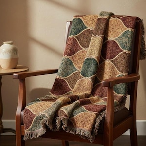 Puede incluir: Una manta tejida con un patrón de formas curvas en tonos marrón, verde y beige, sobre un sillón de madera. La manta tiene bordes con flecos. Un pequeño jarrón decorativo está sobre una mesa auxiliar.