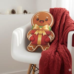 May include: A gingerbread man cushion with a smiling face, holding a red gift box with a gold bow. The cushion is on a white chair next to a red cable-knit throw. Pinecones and candles are in the background, creating a festive scene.