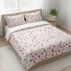 Könnte beinhalten: Ein Doppelbett mit hellem Holzrahmen, bedeckt mit einer weißen Bettdecke und zwei passenden Kissenbezügen. Die Bettwäsche zeigt ein sich wiederholendes Muster aus roten und beigen festlichen Formen, darunter Sterne, Fäustlinge und Schneeflocken.