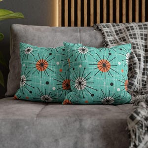 May include: Two square throw pillows with a teal background and a mid-century modern design. The pattern features orange circles, black and white starbursts, and white lines. The pillows are on a gray couch.