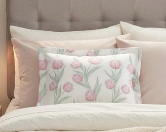 Puede incluir: Una cama con un cabecero tapizado gris claro y varias almohadas. Las almohadas incluyen una funda de almohada blanca con un estampado de tulipanes rosas y fundas de almohada rosas y blancas lisas. La cama está cubierta con un edredón blanco.