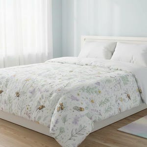 Puede incluir: Funda nórdica blanca con un diseño floral e insectos. El diseño incluye abejas, flores y follaje en tonos amarillos, morados y verdes. La cama está hecha con almohadas blancas y un marco de cama blanco.