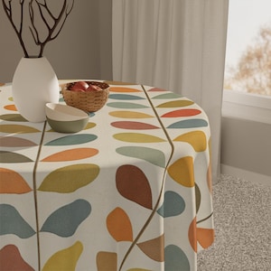 May include: A round tablecloth featuring a cream backdrop and a repeating pattern of colourful leaf shapes in orange, yellow, blue, and brown. A white vase, a small bowl, and a woven basket with fruit are on the table.