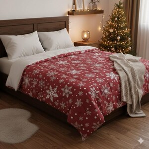 May include: A bed with a red comforter featuring a white snowflake pattern. The bed has white pillows and a light-colored blanket draped over the edge. A Christmas tree with lights is in the background.