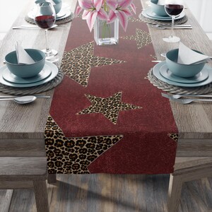 May include: A burgundy table runner with leopard print star designs. The runner is set on a wooden table with place settings, wine glasses, and a vase of pink flowers. The runner adds a decorative touch to the dining table.