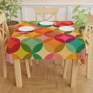 May include: A vibrant tablecloth featuring a geometric design of overlapping circles in red, orange, yellow, green, and pink. The cloth covers a wooden table, with white plates and wooden chairs. The design is reminiscent of the 1960s.