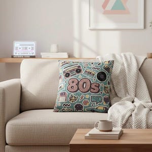 Könnte beinhalten: Ein quadratisches Kissen mit türkisem Hintergrund und einem Retro-80er-Jahre-Thema. Das Kissen zeigt Illustrationen von Kassetten, Boomboxen und das Wort "80s" in Rosa und Braun. Das Kissen liegt auf einem beigen Sofa.