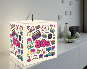 Pode incluir: Uma lâmpada quadrada com fundo creme é decorada com gráficos coloridos com tema dos anos 80. O design inclui cassetes, boomboxes, fones de ouvido e a palavra "80s" em rosa. Uma alça preta arqueia-se por cima.