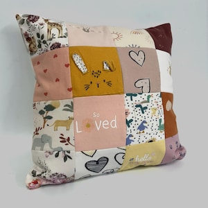 May include: A square patchwork cushion featuring various fabric squares with prints of animals, hearts, and flowers. One mustard yellow square displays embroidered rabbit ears, while a light pink square features the white text So Loved.