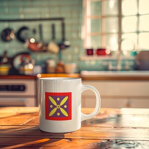 May include: White ceramic mug with a colorful design. The design features a square with a red border, a purple background, and yellow petal-like shapes around a central yellow circle. The mug is on a wooden table in a kitchen setting.