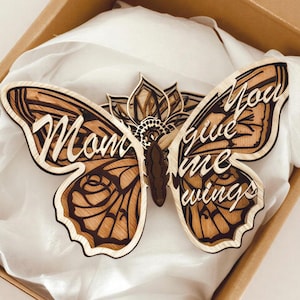 May include: A wooden butterfly with the words "Mom" and "You give me wings" laser-cut into the wings. The butterfly is in a cardboard box with white tissue paper. The butterfly has a floral design in the center.