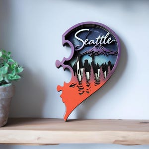 May include: A layered wooden heart-shaped wall art piece. The design features a Seattle cityscape with a mountain and trees, and the word "Seattle" in white script. The color scheme includes purple, blue, orange, and white.