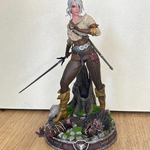 Pode incluir: Estatueta detalhada de uma mulher com cabelo prateado, vestida com calças de couro castanho e uma camisa bege, segurando uma espada. A base apresenta um design de madeira com cabeças de monstros e um emblema de escudo.