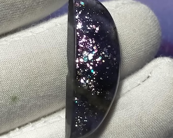 May include: A semi-circular gemstone with a dark purple base and shimmering inclusions. The stone is held by a gloved hand, highlighting its polished surface. The inclusions create a glittery effect, reflecting light. The stone is a cabochon.