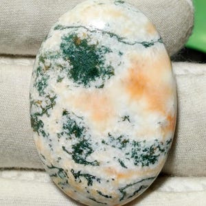 Könnte beinhalten: Oval geformter Edelstein mit polierter Oberfläche. Der Stein zeigt eine Mischung aus Weiß, Grün und Orange. Die grünen Bereiche haben ein moosartiges Aussehen, während die orangefarbenen Abschnitte weich und diffus wirken. Der Stein wird wahrscheinlich für Schmuck oder Dekorationszwecke verwendet.