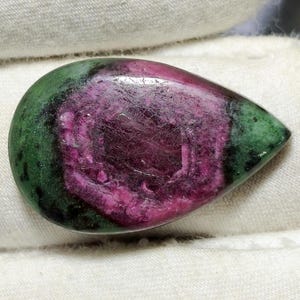 May include: A teardrop-shaped gemstone with a vibrant color combination. The stone features a deep magenta center surrounded by a ring of green and a green border. The polished surface reflects light.