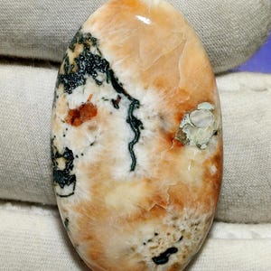 Könnte beinhalten: Oval geschliffener Edelstein mit polierter Oberfläche. Die Farbgebung umfasst Pfirsich, Creme sowie Bereiche in Dunkelgrün und Schwarz. Das Muster erinnert an eine Landschaft mit organischen Formen und Texturen. Der Edelstein ist wahrscheinlich für Schmuck oder Dekorationszwecke bestimmt.