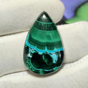 May include: A teardrop-shaped malachite gemstone featuring distinct concentric bands of deep forest green and vibrant turquoise blue. The polished surface displays a smooth, glossy finish with intricate natural patterns and dark green swirls.