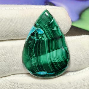 Könnte beinhalten: Ein tropfenförmiger Malachit-Edelstein mit tiefgrünen Bändern und Wirbeln sowie deutlichen Flecken aus leuchtend türkisblauen Mineralieneinschlüssen, poliert zu einer glatten, reflektierenden Oberfläche mit einer klaren, glänzenden Textur.
