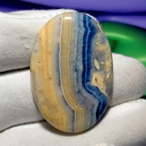 May include: Oval-shaped gemstone with a layered pattern of blue, yellow, and cream colors. The polished surface reveals natural bands and variations, creating a unique and organic appearance. The stone is held by a white surface.