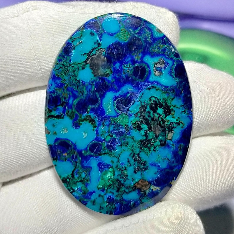 Peut inclure: Pierre pr&eacute;cieuse ovale avec un motif saisissant de bleus profonds, de teintes sarcelle et de noirs. La surface polie pr&eacute;sente un motif naturel et tourbillonnant, cr&eacute;ant un aspect unique et attrayant. La pierre est tenue par une main gant&eacute;e.