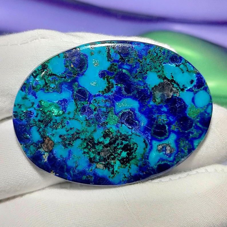 Peut inclure: Pierre pr&eacute;cieuse d'azurite ovale avec une surface polie. La pierre pr&eacute;sente un m&eacute;lange vibrant de teintes bleues et vertes, avec des motifs de veinage bleu fonc&eacute; et noir. Le bijou est pos&eacute; sur un fond en tissu blanc.