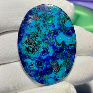 Peut inclure: Pierre pr&eacute;cieuse d'azurite de forme ovale avec une surface polie. La pierre pr&eacute;sente un m&eacute;lange vibrant de teintes bleues, turquoise et vertes, avec des zones plus sombres cr&eacute;ant de la profondeur et de la texture. La pierre est sur fond blanc.
