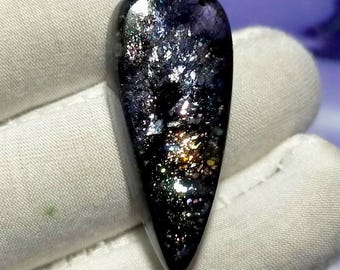 May include: A teardrop-shaped gemstone with a dark, glossy surface. The stone features a speckled pattern of silver, gold, and iridescent flecks, creating a galaxy-like appearance. The gem is held against a white fabric background.
