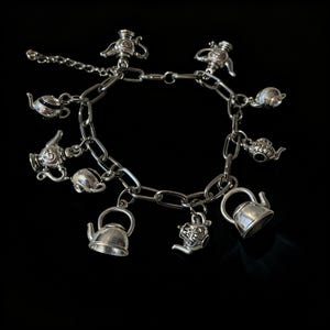 May include: A silver-toned charm bracelet featuring multiple dangling teapot and kettle charms attached to a thick oval link chain. The bracelet includes an adjustable extender chain with a small clasp, showcasing various tea-themed metal ornaments.
