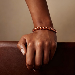 May include: A bracelet composed of a single strand of polished, metallic copper-colored spherical beads. The beads are uniform in size and shape, creating a continuous circular band that rests around the wrist with a smooth, reflective finish.