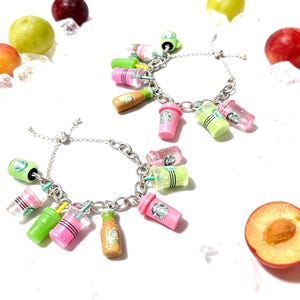 May include: Two silver charm bracelets with colorful miniature drink charms, including pink, green, and clear cups, and brown bottles. The charms are detailed with straws and logos. The bracelets are displayed with fresh fruit on a white background.