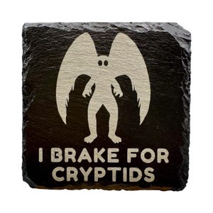 May include: Black square slate coaster with a white graphic of a winged humanoid figure. The text "I BRAKE FOR CRYPTIDS" is below the figure. The coaster has a rough, natural edge.