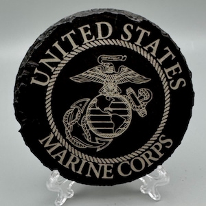 May include: A round, black slate coaster featuring the United States Marine Corps emblem. The emblem includes an eagle, globe, and anchor design, with the words "United States Marine Corps" in a white, rope-style border.