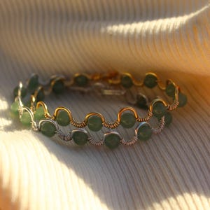 May include: Two bracelets featuring green beads and gold and silver wirework. They are displayed on a cream-coloured fabric, with sunlight creating a warm, golden glow.