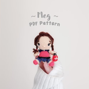 May include: A small crocheted doll with brown hair in pigtails, wearing a pink top and blue shorts, holding a red bag with a white cross. The text above the doll reads ~ Meg ~ PDF Pattern. The doll features black eyes and pink shoes.