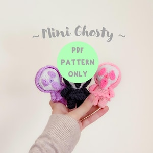 Peut inclure: Trois figurines de fantômes crochetés en violet, noir et rose, chacune avec un visage et une capuche blancs. Le texte "Mini Ghosty" et "PDF Pattern Only" sont visibles. Les figurines sont tenues dans une main sur fond clair.